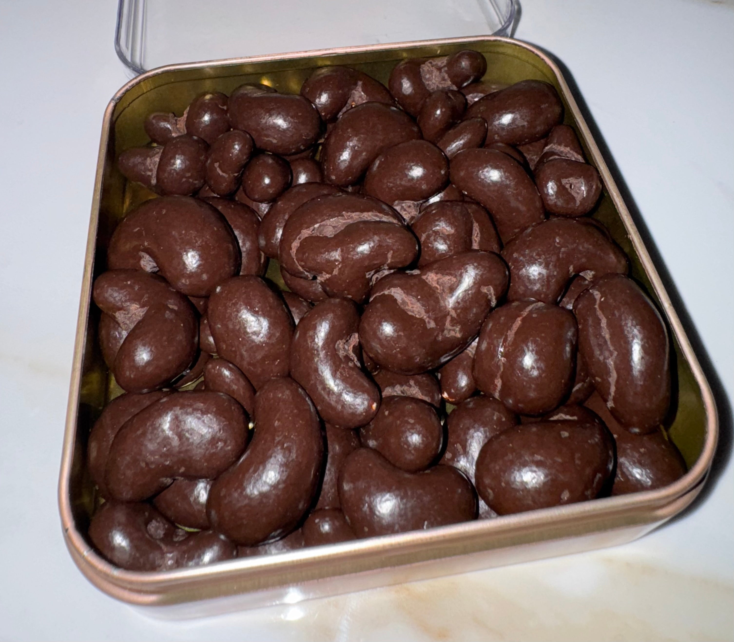 A close-up shot of glossy chocolate-covered cashews piled in a square metal tin with a clear lid, set on a light-colored surface.